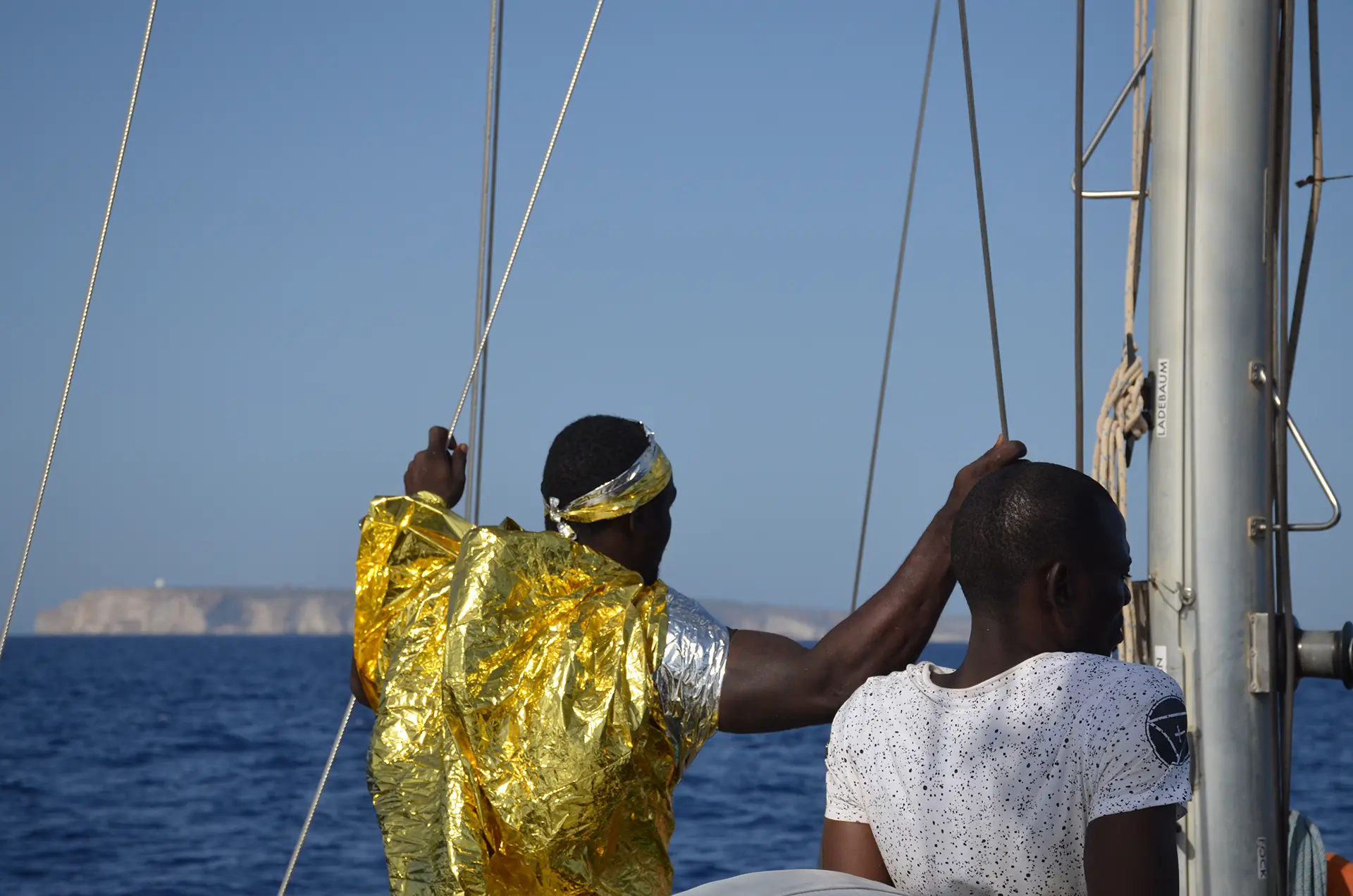 Zwei gerettete Menschen an Deck, eine in Rettungsdecke, blicken auf die Küste